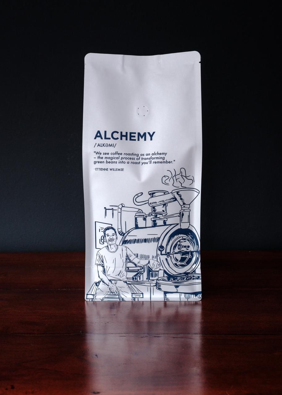 White coffee bag with 'Alchemy' branding on a dark background. Premium coffee beans with a glossy roast, crafted for coffee lovers who savor every sip